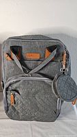 Front view of grey diaper bag showing multiple compartments, quilted front pocket, leather zipper pulls, and attached round pouch.
