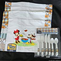 Three Disney Mickey Mouse placemats featuring a cooking scene with Minnie and Mickey, bordered by plaid and floral designs, displayed alongside the flatware set still in packaging.