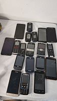 Collection of 16 assorted cellphones including flip phones, smartphones, and a Blackberry style phone, all with noticeable wear and damage.