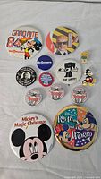 Top down photo of assortment of 12 vintage and modern buttons showing Disney themes, political, and novelty designs.