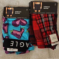 Two pairs of new American Eagle stretch boxers, size medium, one with eggplant print and one red plaid, showing waistband and packaging.