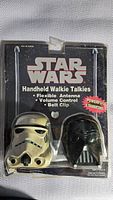 Front view of Star Wars branded packaging containing two handheld walkie talkies shaped like Stormtrooper and Darth Vader helmets.