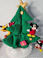 Front view of green Santa hat shaped like Christmas tree with plush Disney characters Minnie Mouse, Mickey Mouse, and Pluto attached around the tree branches. Yellow star topper and white faux fur trim at base.