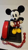 Photo of full Mickey Mouse telephone showing figure standing adjacent to red handset and coiled cord on black base.