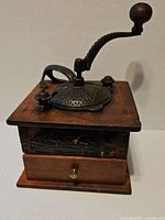 Side perspective showing the cast iron top with embossed decoration, twisted handle with wooden knob, wooden box base, and small front drawer with brass knob showing wear.