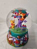 Front view showing Pooh reaching for fruit on purple tree inside snowglobe, Tigger lounging on branch, and blue basket with red fruits on green glitter base
