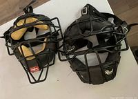 Two black Rawlings baseball/softball catchers masks with wire face guards and tan padding inside, laid side by side on wooden floor.
