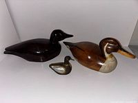 Photo showing three duck decoys including a large dark wood carved duck, a wooden pintail duck decoy with painted details, and a small decorative brass and mother of pearl duck.