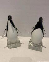 Front view of the pair of glass penguin figurines on clear square bases, showing black and white coloring and stylized shape.