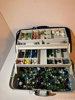 Photo showing the tackle box opened to reveal several compartments filled with marbles of different colors and sizes, with larger marbles stored on the bottom.