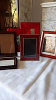 Photo showing front of seven assorted photo frames on a white patterned cloth, including red, brown, and wooden frames with different designs.