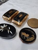 View of two rectangular clothes brushes with black tops decorated with gold swirls and central blank shield, gold metal sides and bristle backs, plus three powder compacts including one with painted boat and landscape scene and another with white floral design and one gold textured lid compact