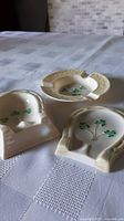 Three Belleek porcelain items on tablecloth: two shamrock-decorated spoon rests and one ashtray