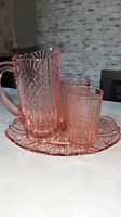 Photo showing the pink Depression glass jug, two tumblers, and matching tray on a table, highlighting the overall set with visible geometric patterns.
