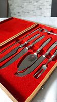 Set of six chrome and steel entertainment utensils arranged inside red velvet-lined wooden box.