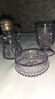 Three pieces of amethyst pressed glass: syrup pitcher with metal lid, pedestal bowl, and diamond point bowl.