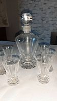 Photo showing the crystal decanter with round stopper surrounded by six matching shot glasses, all with hexagonal bases and clear crystal material.