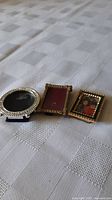 Three vintage miniature portrait frames displayed on a white textured cloth; one round silver frame and two rectangular gold frames, one with photo insert.