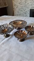 Four vintage silverplated bowls and candle holders on a white cloth with intricate filigree work visible.