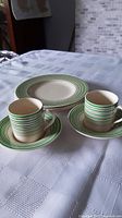 Photo showing 2 espresso cups with handles on matching saucers and 3 matching side plates, all with green crayon lines pattern.