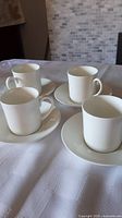 Four white bone china cups with matching saucers set on a white tablecloth, background tile wall visible.