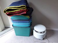 Stacked Rubbermaid totes with folded moving blankets and floor fan beside