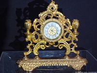 Front view of gilt metal mantel clock showing dial, scrollwork and candle holders