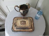 Top view of silver-plated pitcher and lidded serving dish on table for full lot context