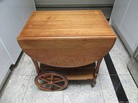 Full view of tea cart with leaves half raised showing carved motif, wheels, drawer, lower shelf