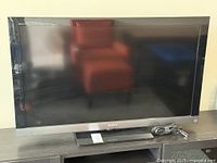 Front view of Sony BRAVIA TV on black stand with power cord coiled beside it, screen reflects surroundings.