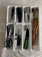White plastic drawer organizer holding wooden spoons, scissors, peeler, melon baller, thermometer, and other utensils.