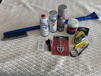 Photo of automobile care tools on white surface showing ice scraper with brush, paint spray cans, body filler, battery and air pressure sensors, sanding sponge, and gauge.