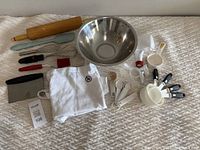 Full view of all items including stainless steel bowl, measuring cups & spoons, piping bag, egg beaters, rolling pin, spatula, dough scraper, brush, and apron.