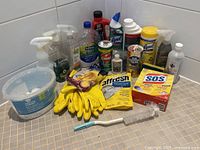 Full overview of assorted bathroom and kitchen cleaning supplies arranged on a tiled surface, showing gloves, bottles, and cleaning pads.