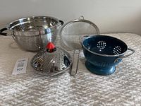Photo of all four items together: large stainless steel colander, blue colander, stainless steel strainer, and collapsible steamer with red knob