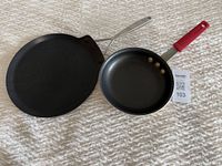 Henckels crepe pan and Tramontina 8-inch frying pan side by side on a textured surface, showing the overall appearance and features of both pans.