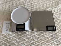 Photo of two digital kitchen scales side by side on a textured surface, showing one scale with a white round platform and the other with a stainless steel weighing surface.