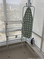 Photo showing collapsed drying rack with multiple horizontal bars, ironing board standing upright with decorative cover, and T-Fal Ultraglide Pro iron on floor.