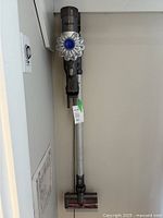 Dyson V6 Origin vacuum cleaner mounted on wall dock in storage position. Main handheld vacuum unit with long extension wand and motorized floor head attachment shown.