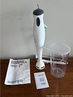 Hand blender with cup, lid, and instruction manual laid on surface, showing overall contents of the lot
