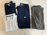 Two long sleeve Helly Hansen shirts (white and navy) and two pairs of Helly Hansen pants (navy cargo pants and moss lightweight pants) arranged flat and visible in full.