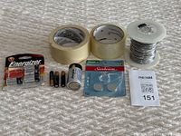 Photo showing masking tape, packing tape, aluminum wire spool, assorted tested batteries including 9V, AA, AAA, C, D sizes, and button cells on a bed spread.