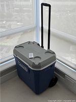 Full exterior view of the Coleman large cooler showing blue body, gray lid with cup holders, telescoping handle extended, and one visible wheel.