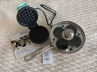 Photo showing the mini turquoise Dash waffle maker open next to the Demeyere stainless steel egg poacher pan with glass lid on a textured fabric surface.