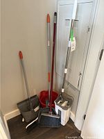 Photo showing two mop buckets (red and white with wringer), three brooms (various handle colors), two dust pans (black and white), and one mop leaning against a wall near electrical panel.