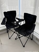 Two black portable camping chairs set up side by side on a tiled floor near a window, showing fabric seats, backs, armrests, and cup holders on each chair's right armrest.