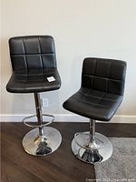 Two black vinyl bar stools with backrests, chrome bases, square stitching pattern on seats and backs.