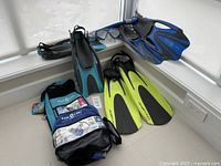 Photo shows three sets of snorkeling equipment including dark blue and light blue fins, yellow fins, two travel bags, and snorkel masks and snorkels arranged on a corner floor space.