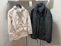 Front view of two women's jackets hanging side by side on hangers: an off-white spring jacket and a dark green insulated winter jacket.