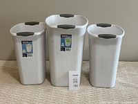 Photo showing three white Sterilite garbage bins placed side by side on the floor, two smaller bins on either side and a taller one in the center.